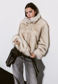 Faux fur jacket in cream with a high collar, wide sleeves, and a fluffy texture, paired with dark high-waisted jeans and a leather belt.