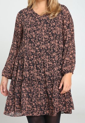 Kvinde iført en sort kjole med pink blomsterprint og lange ærmer, stående mod en ensfarvet lys baggrund.