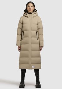 Sieviete valkā garu beidzamo pelēkā ziemas mēteli ar kapuci, melnas legingi un melnas šņorējamas zābaki, stāvot vērsta uz priekšu.