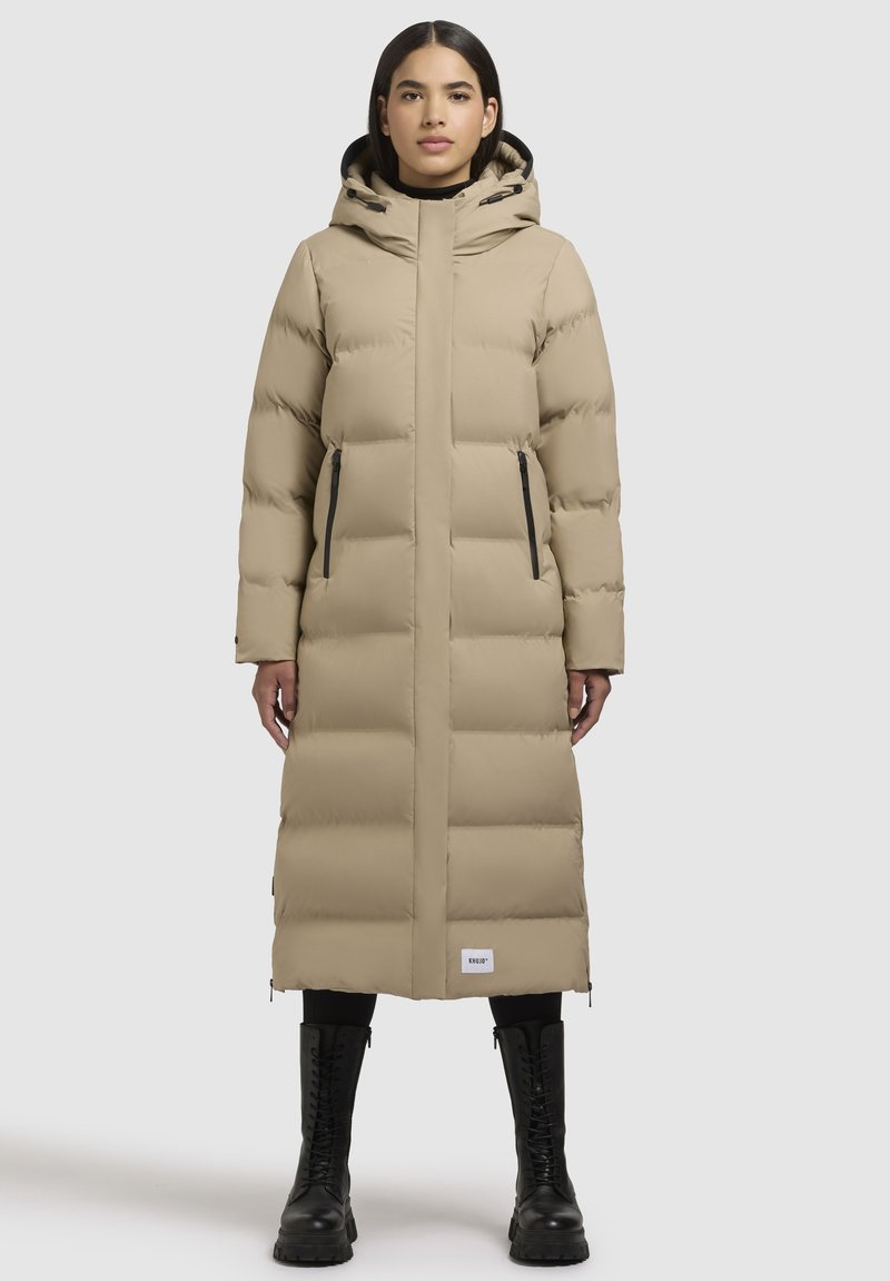 Sieviete valkā garu beidzamo pelēkā ziemas mēteli ar kapuci, melnas legingi un melnas šņorējamas zābaki, stāvot vērsta uz priekšu.