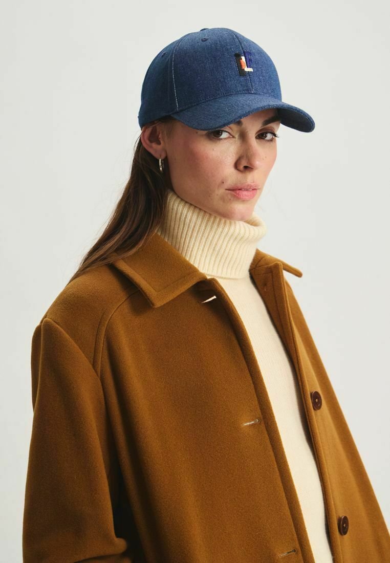 Casquette de baseball en denim bleu avec un logo en relief, portée avec un manteau beige et un col roulé crème. Elle se distingue par une visière courbée et des coutures classiques.