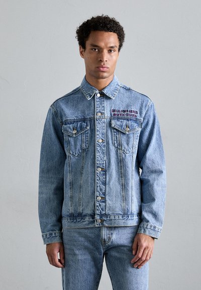 Veste en denim bleu clair avec un col, poches avant et texte brodé sur le côté gauche de la poitrine. Comprend des boutons métalliques et des coutures surpiquées.
