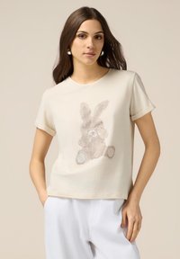 T-shirt crème à manches courtes avec un design de lapin texturé dans des tons marron clair et beige, présentant une coupe décontractée et un tissu doux.