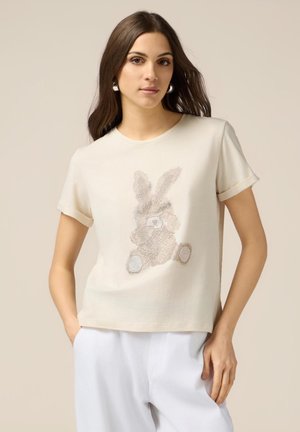 Cremfarbene Kurzarm-T-Shirt mit einem strukturierten Hasendesign in hellbraunen und beigen Tönen, das einen lässigen Schnitt und einen weichen Stoff aufweist.