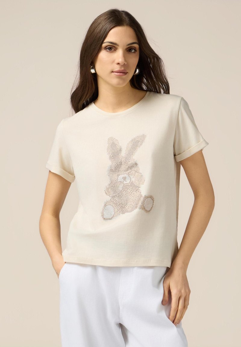 T-shirt crème à manches courtes avec un design de lapin texturé dans des tons marron clair et beige, présentant une coupe décontractée et un tissu doux.
