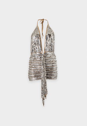Femme portant une mini-robe dos nu à sequins argentés avec un décolleté en V profond et des talons hauts noirs, debout devant un fond clair uni.