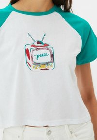 Kratký rukáv, zkrácený tričko s bílým tělem a tyrkysovými rukávy. Obsahuje barevnou vyšívanou retro grafiku televizoru se slovem "mí peace".