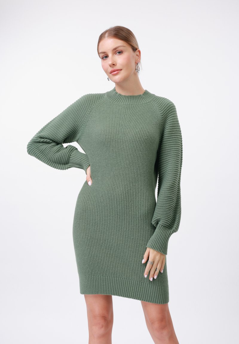 Abito in maglia di colore verde oliva, con texture a costine, maniche lunghe e puffate, e scollo rotondo. Silhouette aderente che termina sopra il ginocchio.