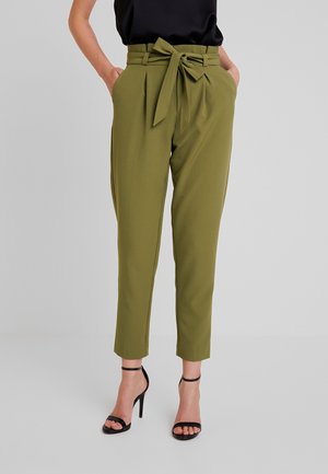 Mujer vistiendo pantalones caqui de talle alto y corte cónico con cinturón atado y sandalias negras de tacón con tiras en el tobillo, con las manos en los bolsillos.