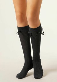 Beine mit dunkelgrauen, gestreiften knielangen Socken, die außen kleine schwarze Satinschleifen haben, vor neutralem Hintergrund.