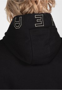 Sudadera negra con capucha que presenta una etiqueta bordada en contraste en la parte superior. Tejido suave con un corte estándar y un escote relajado.