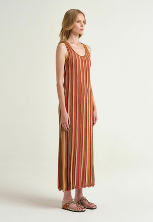 Donna in piedi con un abito lungo senza maniche a righe verticali multicolori, indossa sandali aperti con motivo pitone su uno sfondo uniforme.