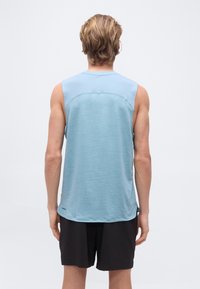 Άνδρας με ξανθά μαλλιά, φορώντας αμάνικη αθλητική μπλούζα ανοιχτού γαλάζιου χρώματος και μαύρο σορτς, γυρισμένος με την πλάτη προς το φωτεινό μονόχρωμο φόντο.