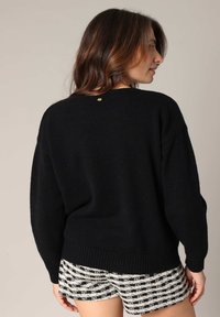 Pull en tricot noir avec un col rond, coupe loose et ourlet côtelé. Un petit accent doré est visible à l'arrière près du col.