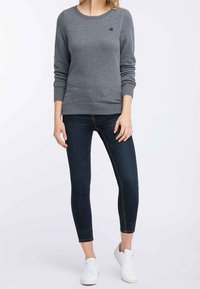 Sudadera gris con puños y dobladillo acanalados, combinada con jeans skinny oscuros y zapatillas blancas. Detalle del logotipo en el pecho.