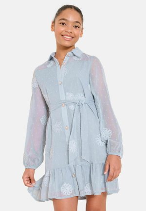 Mosolygó lány világoskék, áttetsző ruhában, virágos hímzéssel, gombos elejű, deréknál megkötős, fodros szegéllyel, fehér háttér előtt.