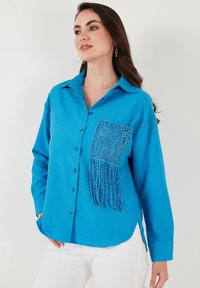 Camicia button-up blu brillante in tessuto testurizzato, con tasca in pizzo e dettagli con frange. Maniche lunghe e vestibilità rilassata.