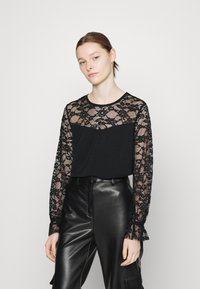 Top preto com mangas compridas em rendas que apresentam padrões florais, combinado com calças de couro pretas de cintura alta. Decote redondo simples, corte relaxado.