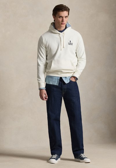 Νέος άντρας που φοράει ένα εκρού hoodie Polo Ralph Lauren πάνω από ένα μπλε τζιν πουκάμισο, σκούρα τζιν και ναυτικά αθλητικά παπούτσια με λευκές σόλες.