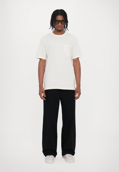 Vit bomulls t-shirt med en bröstficka, i kombination med löst sittande svarta byxor och ljusa sneakers. Minimalistisk design och avslappnad stil.