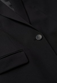 Mustapuku takki, jossa on välikärki, yksi musta nappi kiinnitettynä ja pahvikotelo näkyvissä vasemmalla puolella.