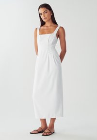 Žena v bielej bezrukávovej midi šaty a čiernych plochých sandálach stojí na pozadí bielej farby.