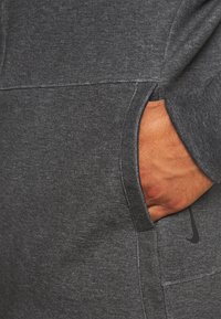 Main reposant à l'intérieur de la poche avant d'un sweat-shirt Nike gris foncé avec des coutures visibles et un logo près du bord de la poche.