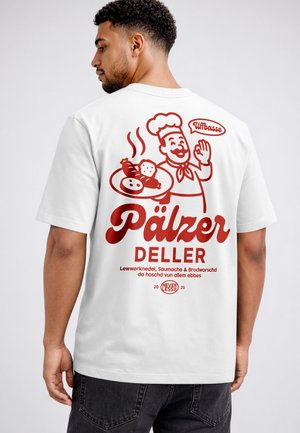 Mann trägt weißes T-Shirt mit rotem Motiv eines lächelnden Kochs, der einen Teller mit traditionellem deutschem Essen hält, und deutschem Text auf dem Rücken.