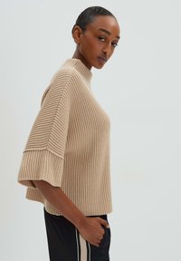 Beige gestrickter Pullover mit hohem Kragen, weiten kurzen Ärmeln und Rippstruktur, kombiniert mit schwarzen Hosen mit seitlichen Streifen.