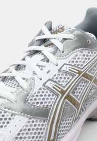 Chaussure de sport blanche avec tige en mesh et accents argentés. Présente un col rembourré gris et des lacets blancs. Logo Asics visible sur le côté.