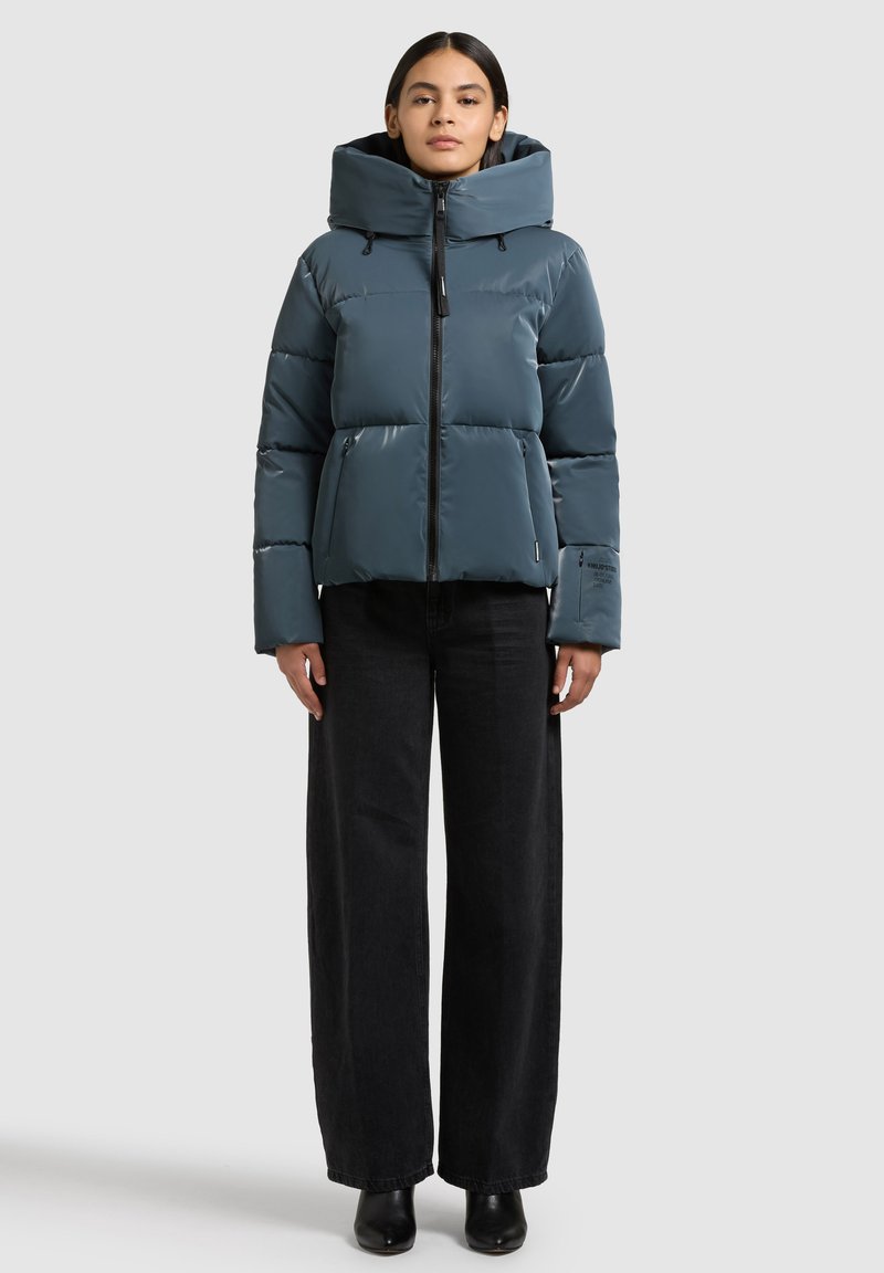 Pūkinė striukė subtilios žalsvos mėlynos spalvos, su aukšta apykakle, užtrauktuku ir šoninėmis kišenėmis. Lygus, blizgus paviršius su šiek tiek stambiu siluetu.