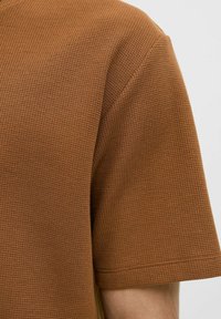 Camisa marrón de manga corta hecha de tejido texturizado de punto gofre. Presenta un corte relajado con un escote redondeado y un patrón sutil.