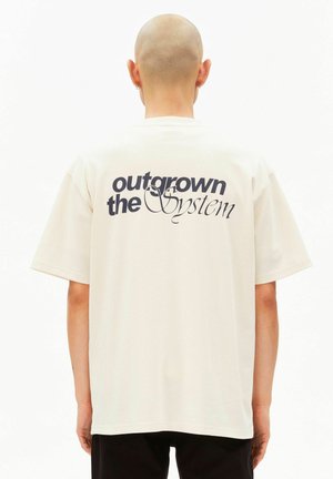 Beiges oversized T-Shirt mit kurzen Ärmeln, auf der Rückseite mit der Aufschrift "outgrown the System" in Dunkelblau bedruckt. Weiche Baumwollstruktur.