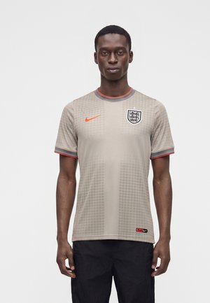 Mężczyzna stojący, ubrany w beżową koszulkę piłkarską Anglii z pomarańczowym logo Nike i herbem, w połączeniu z czarnymi spodniami, na jednolitym białym tle.