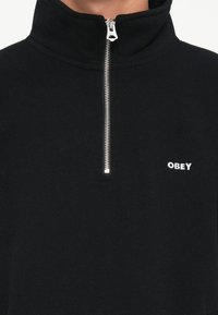 Czarny polarowy pulower pół-zamek z wysokim kołnierzem, z wytrzymałym srebrnym zamkiem błyskawicznym oraz białym logo "OBEY" na lewej piersi. Miękka tekstura.