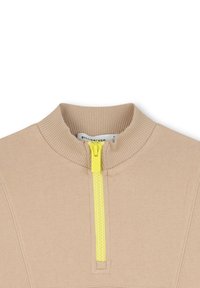 Beige gestricktes Kleidungsstück mit einem gerippten hohen Kragen und einem kurzen gelben Reißverschluss in der Mitte vorne, der ein teilweise weißes Etikett im Kragen zeigt.