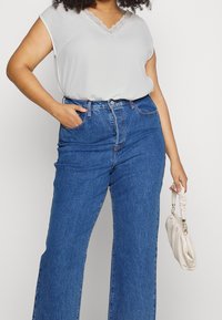 Person med krøllet hår iført en hvid top med blonder og højtaljede blå jeans, som holder en lille hvid rynket håndtaske i højre hånd.