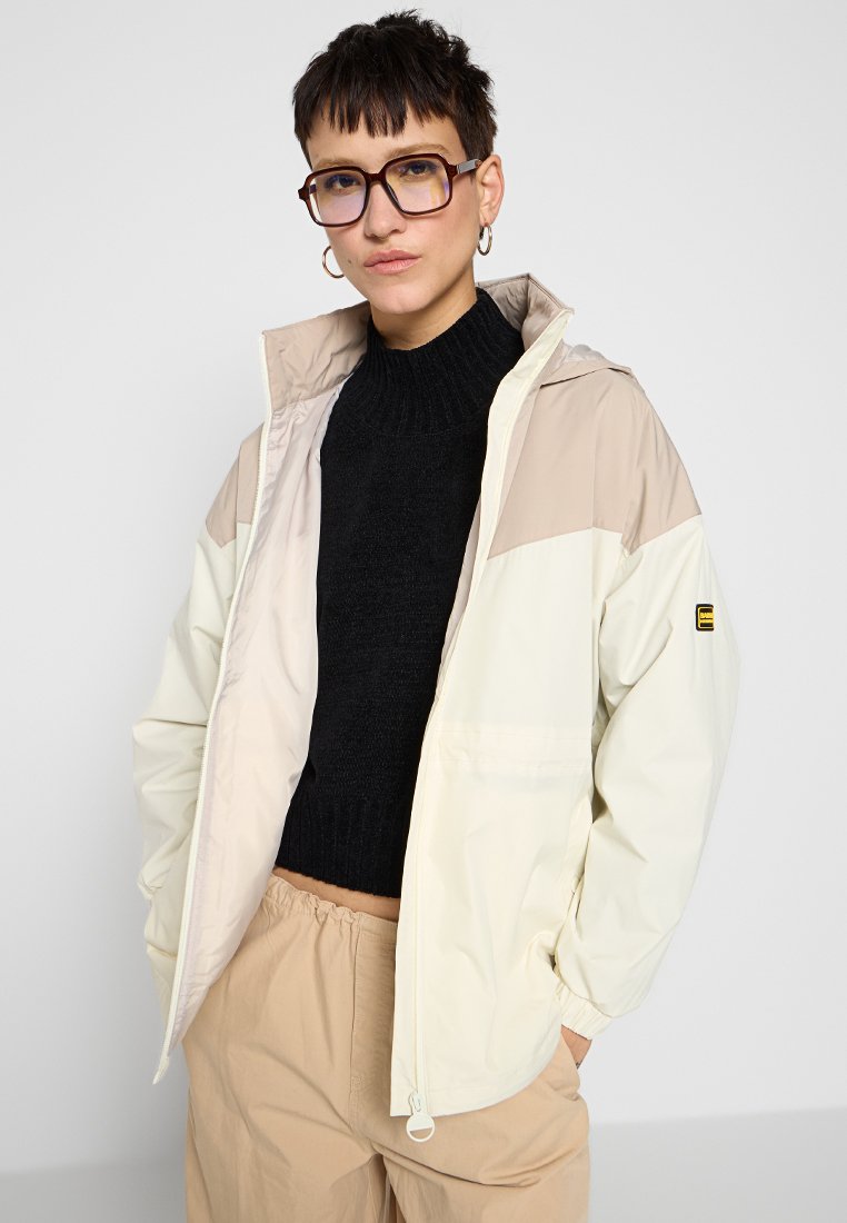 Giacca leggera color blocco crema e beige con zip frontale, cappuccio e due tasche laterali, abbinata a un maglione nero in maglia.