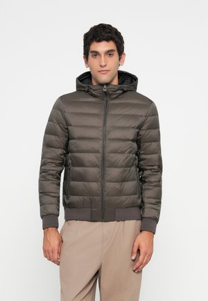 Man wearing a dark gray padded jacket with a hood and beige pants, standing against a plain light background.