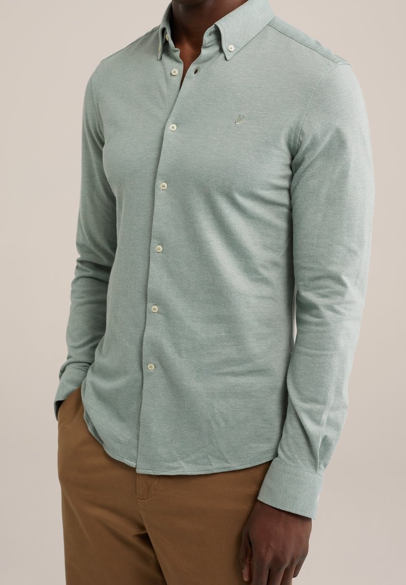 Man som bär en ljusgrön knäppt långärmad skjorta med krage och bruna byxor, står med en hand i fickan.