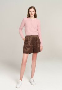Rosa gestrickte Strickjacke mit langen Ärmeln und Knöpfen, kombiniert mit braunen gefalteten Shorts. Weiße Turnschuhe runden das Outfit ab.