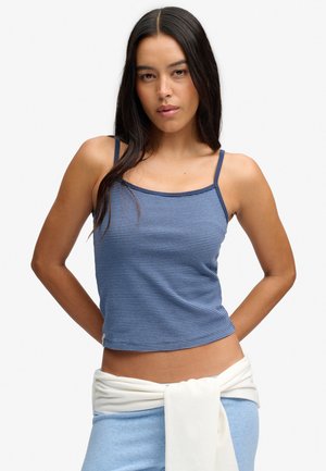 Kvinde med langt mørkt hår iført en tætsiddende blåstribet tanktop og lyseblå bukser med en hvid sweater bundet om livet.