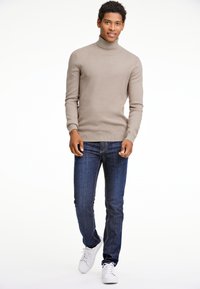 Beige Rollkragenpullover aus geripptem Strickmaterial, kombiniert mit dunkelblauen Jeans und weißen Sneakers, im taillierten Design.