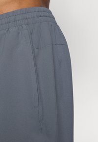 Närbild av en persons midja iklädd mörkgrå byxor med elastiskt midjeband och en sidficka mot en enhetlig ljus bakgrund.