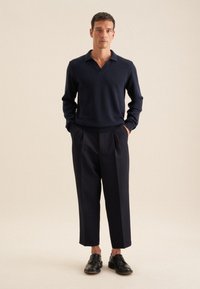 Marineblauer Strickpullover mit V-Ausschnitt, kombiniert mit hochgeschnittenen, gefalteten marineblauen Hosen und schwarzen Lederschuhen, die eine glatte Textur aufweisen.