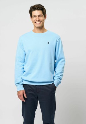 Lys blå crewneck sweatshirt med ribbede manchetter og kant, med et lille sort logo på brystet, parret med mørke bukser.