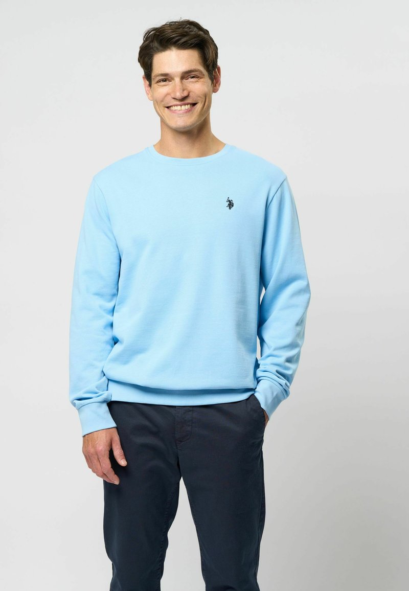 Šviesiai mėlynas crewneck tipo švarkas su raiščiais ant rankovių ir apykaklės, su mažo juodo logotipo detalėmis krūtinėje, derinamas su tamsiomis kelnėmis.