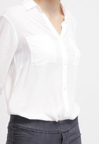 Blusa bianca con bottoni e colletto, con due tasche sul petto e un tessuto morbido e leggero. Indossata con pantaloni grigio scuro.