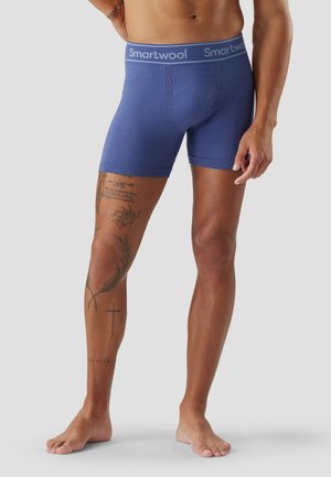 Mandligt overkrop og ben iført blå Smartwool boksershorts, der viser tatoveringer på venstre ben, stående barfodet på lysegrå baggrund.