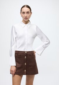 White long-sleeve blouse with a collar, textured fabric, and buttoned front; paired with a brown suede mini skirt featuring gold buttons and pockets.